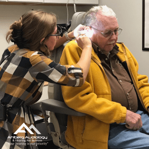 Dr. Kelsey Brittingham conducting ear wax removal with patient, gray-haired man with mustard yellow jacket and brown button-down shirt.